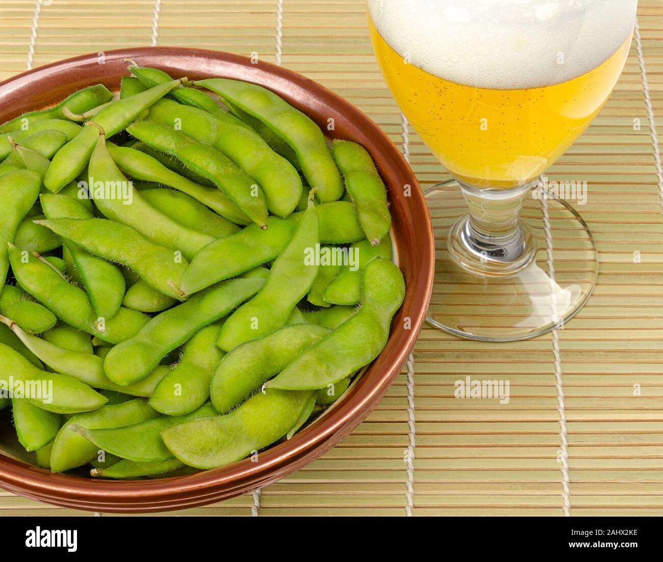 Edamame bolliti in una ciotola con un bicchiere di birra sul tappetino di bambù. Fagioli di soia verde, maodou, cotto in acqua salata. Piatto di lato e gustosi snack. I fagioli di soia. Foto Stock