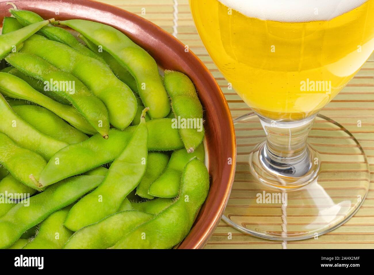 Edamame bolliti in marrone ciotola con un bicchiere di birra sul tappetino di bambù. Fagioli di soia verde, maodou, cotto in acqua salata. Piatto di lato e gustosi snack. I fagioli di soia. Foto Stock
