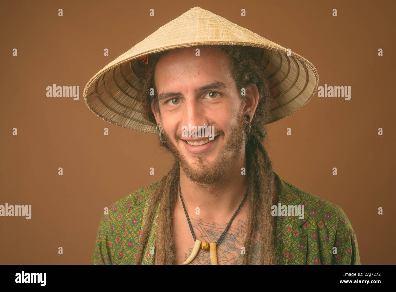 Studio shot di giovani bello uomo ispanico con dreadlocks indossando stile asiatico cappello conico contro sfondo marrone Foto Stock