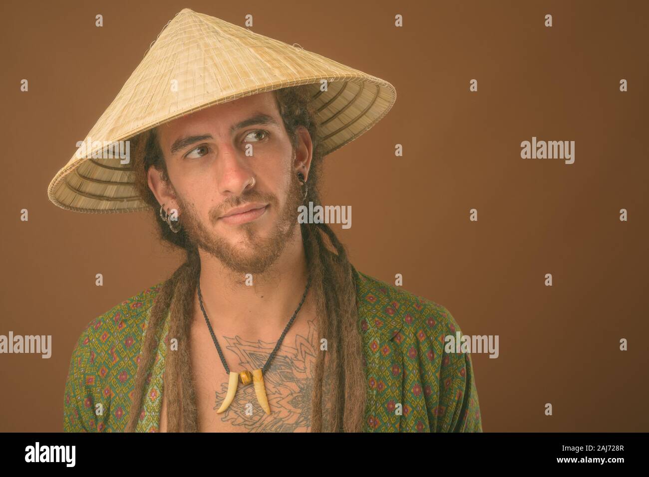 Studio shot di giovani bello uomo ispanico con dreadlocks indossando stile asiatico cappello conico contro sfondo marrone Foto Stock