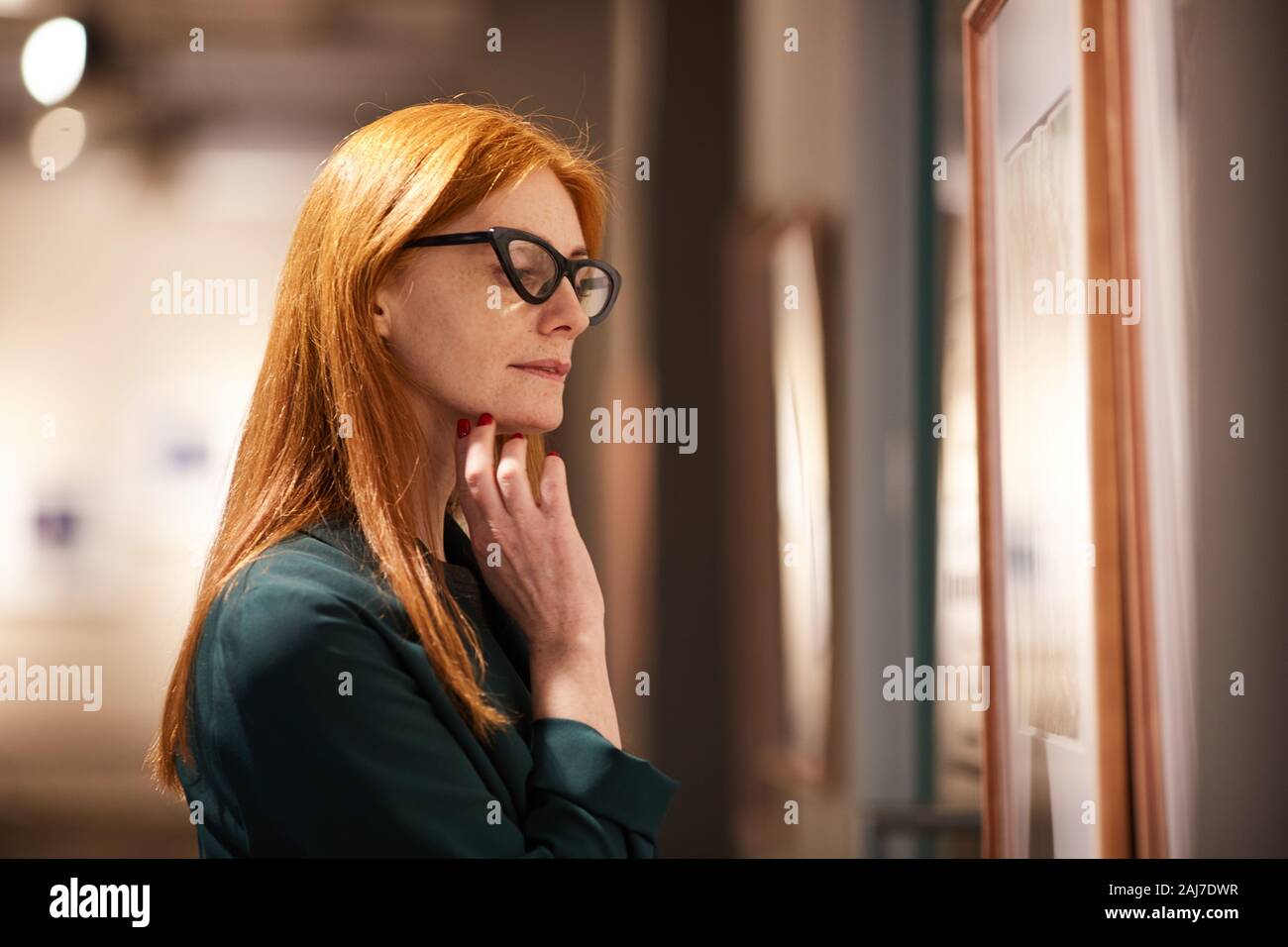 Rosso giovane donna dai capelli in occhiali guardando la pittura e godendo della tecnica nella galleria Foto Stock