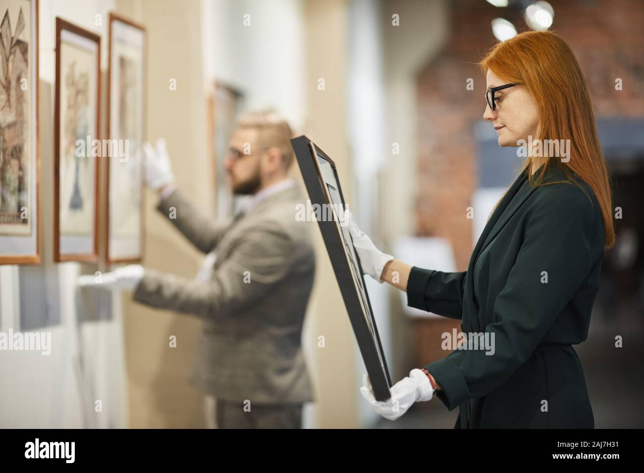 Vista laterale di dai capelli rossi ragazza in occhiali esaminando la pittura nella sua mano e sceglierne uno per se stessa con uomo al lavoro Foto Stock