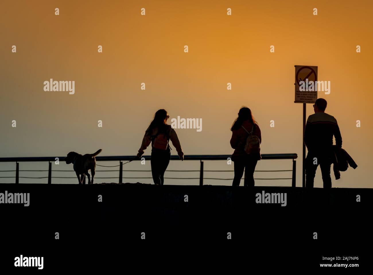 Le persone sul lungomare al tramonto passeggiando a piedi il cane nella luce arancione del tramonto sulla costa atlantica in Portogallo. Fotografia: Tony Taylor Foto Stock