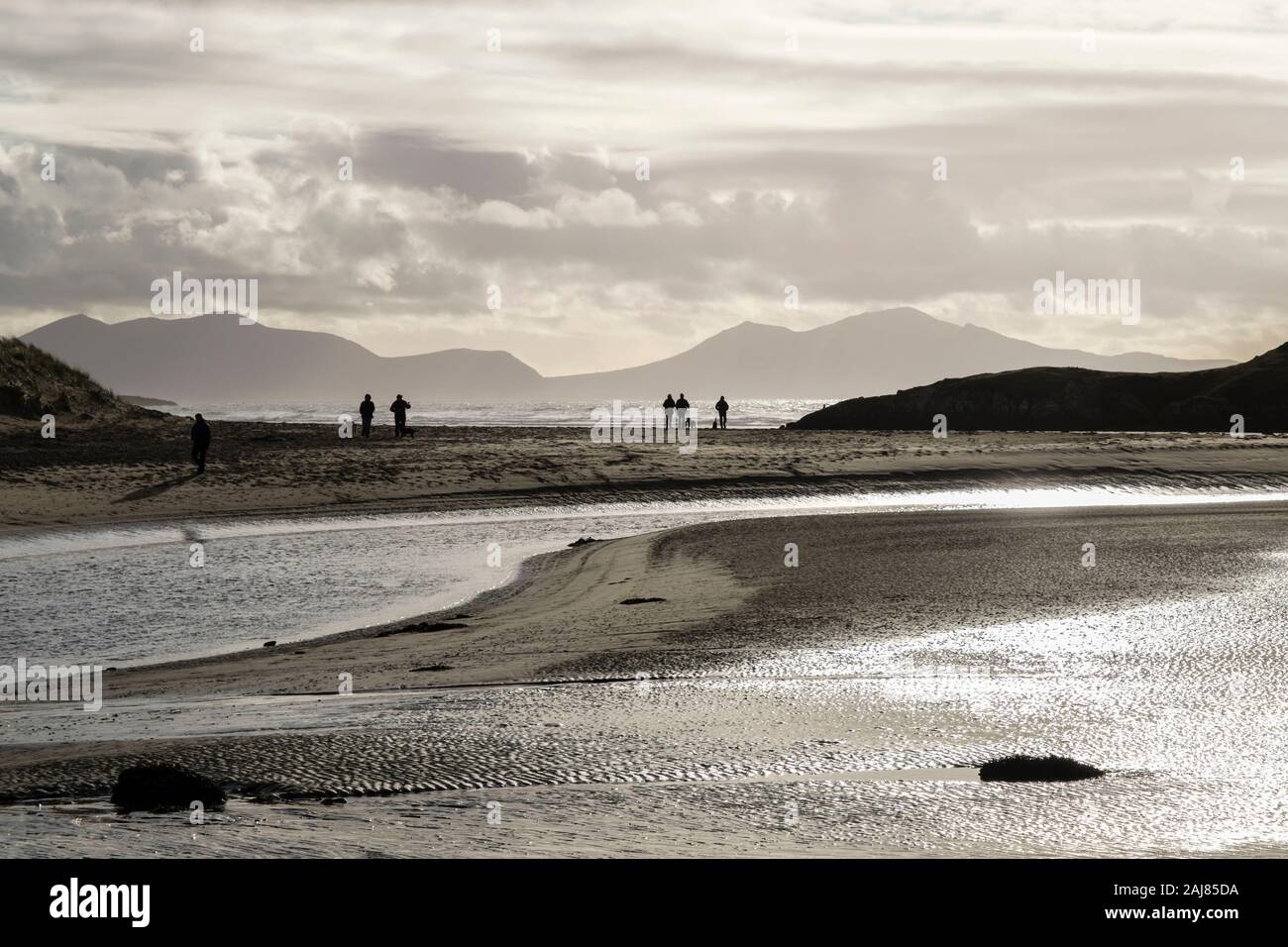 Vista retroilluminato attraverso Afon Ffraw fiume con la gente che camminava sul Traeth Mawr spiaggia con montagne in inverno. Aberffraw Isola di Anglesey nel Galles REGNO UNITO Foto Stock