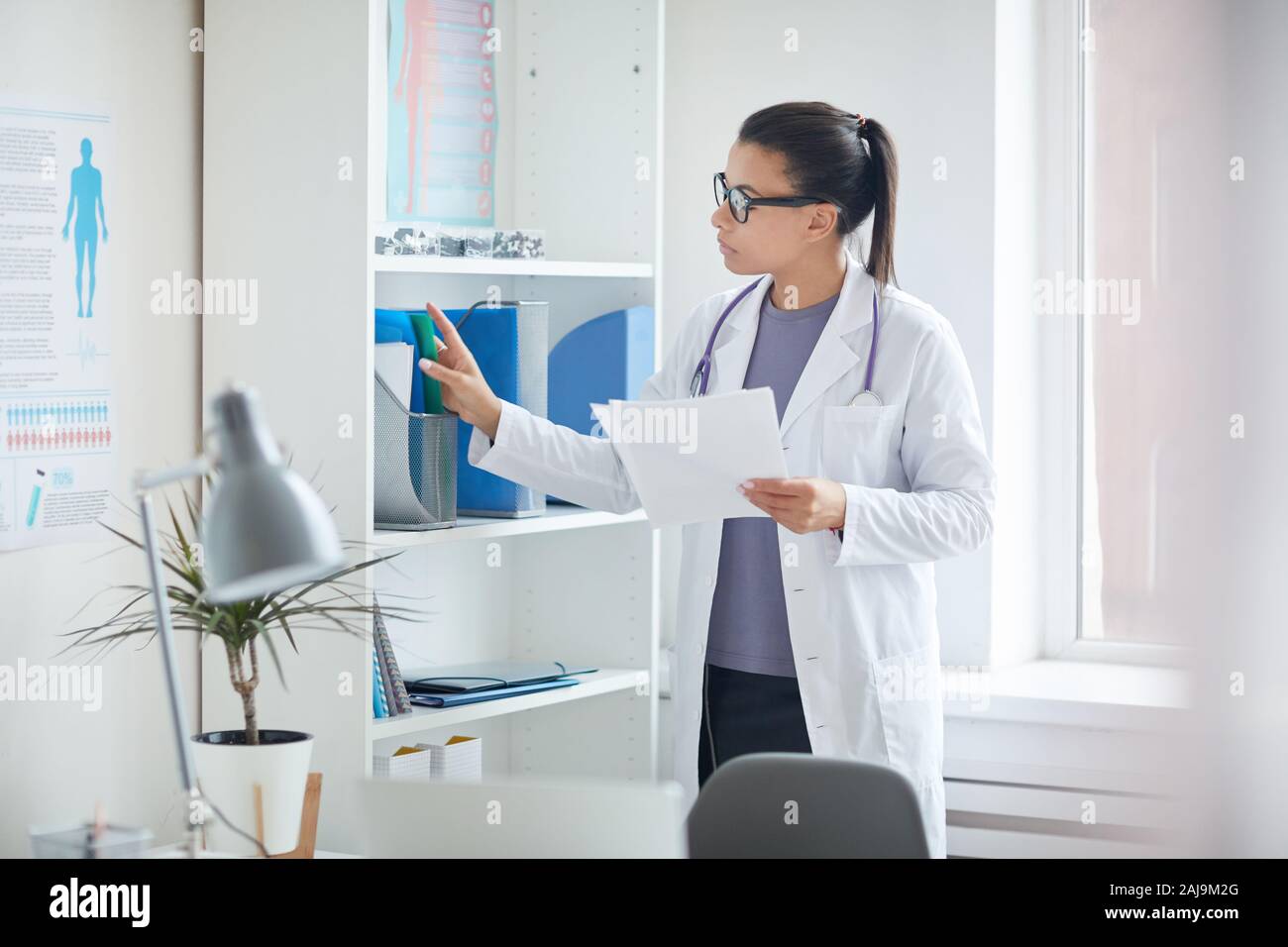 Medico donna in camice bianco in piedi vicino alla libreria e cercando le schede mediche dei suoi pazienti alla clinica medica Foto Stock