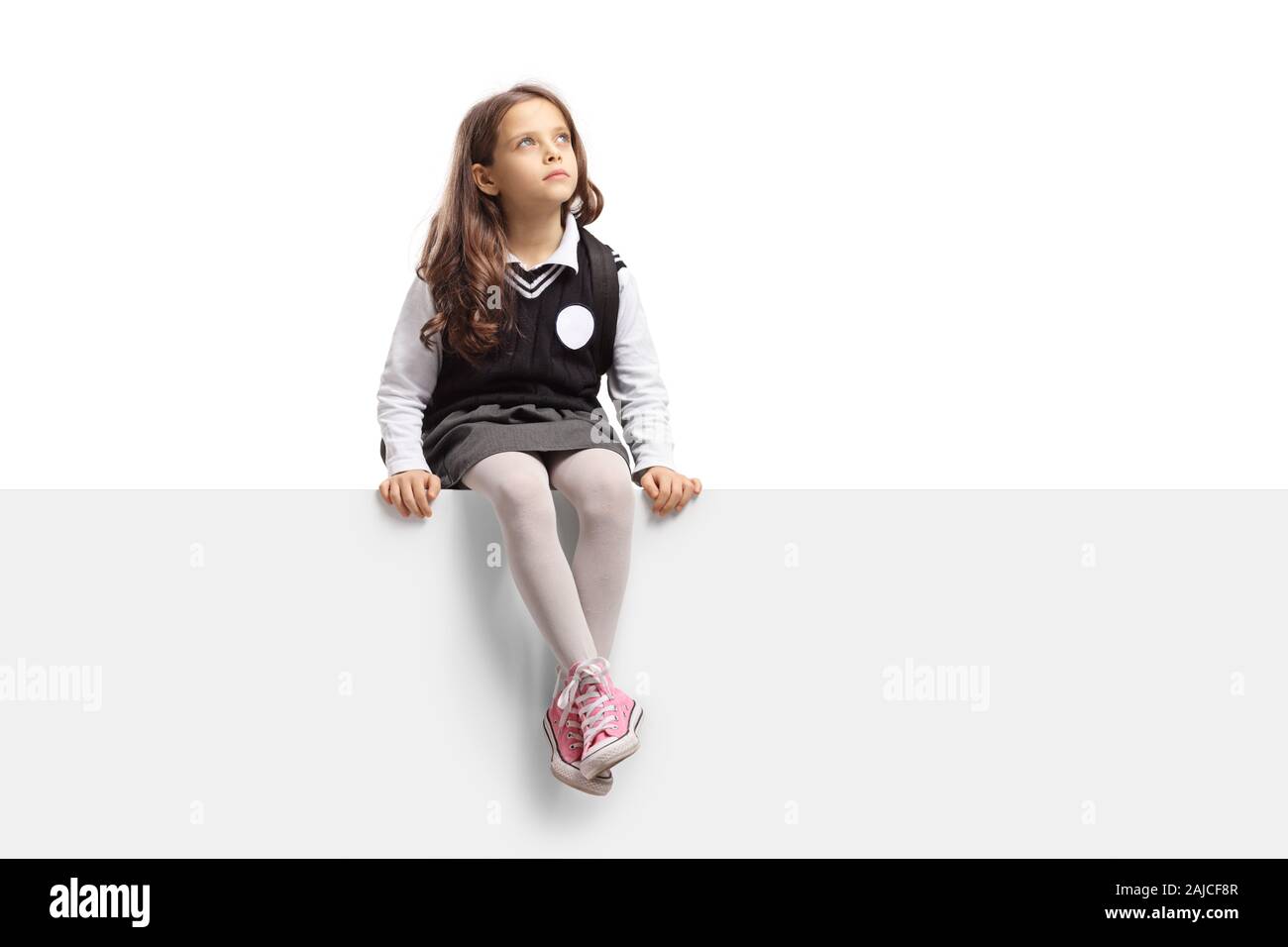 Schoolgirl in uniforme seduto su un pannello vuoto e guardando in alto isolato su sfondo bianco Foto Stock
