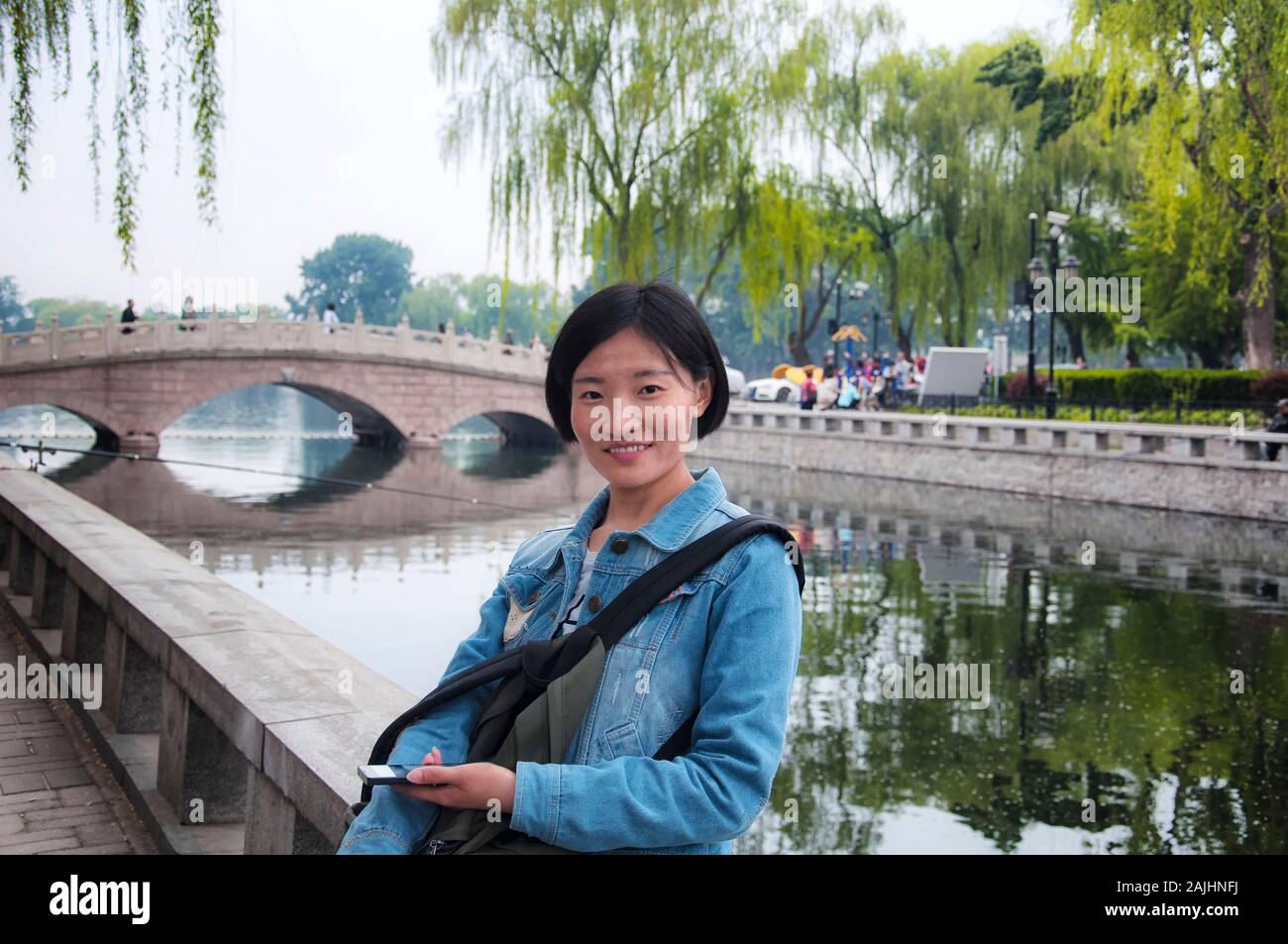 Una donna cinese in possesso di una smart phone appoggiato contro una parete con Jinding ponte sopra il lago Qianhai negli Hutong scenic area in background in Pechino Foto Stock