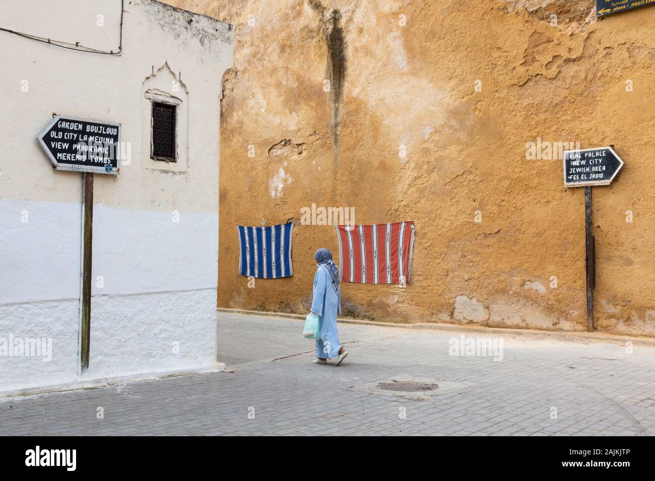 Donna in abito blu e con velo sulla testa, segnaletica che mostra il percorso per le attrazioni turistiche e tessuti appesi al muro, Fes (Fez), Marocco Foto Stock