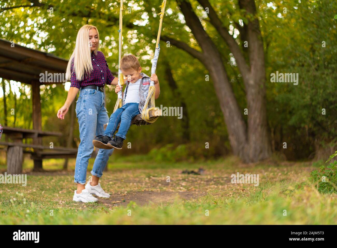 Giovane biondo mom scuote il suo piccolo figlio su altalena in un parco verde. Infanzia felice Foto Stock