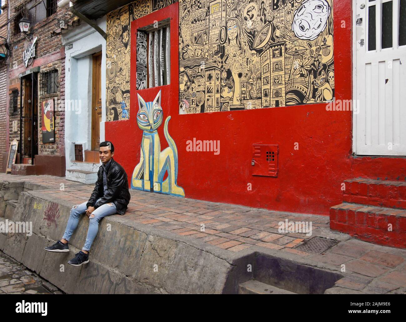 Giovane uomo seduto sulla parete esterna edificio colorato su Calle del Embudo decorate con arte di strada, La Candelaria distretto di Bogotà, Colombia Foto Stock