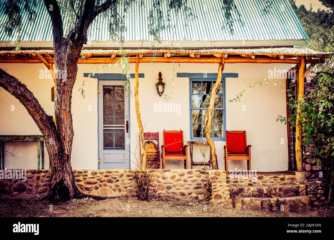 Un albero di Mesquite vicino al palo e albero post portico di una influenza spagnola casa adobe con sedie rosse sul portico in città artigianale di Tubac, AZ, Stati Uniti d'America Foto Stock