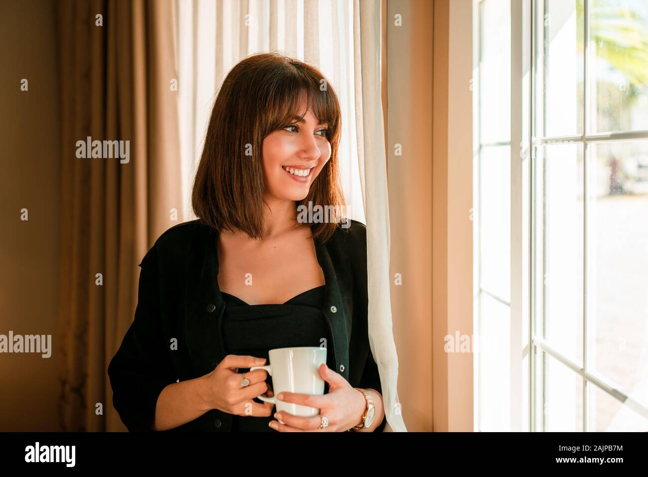 Giovane donna sorridente e bere qualche bevanda e guardando fuori dalla finestra. Foto Stock
