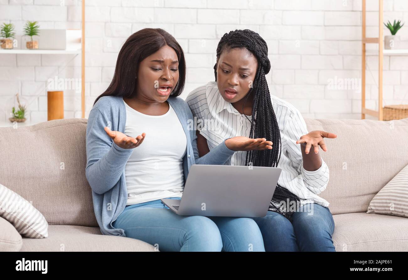 Nessuna connessione a internet. Due deluso afro fidanzate di fronte a problemi con il computer portatile durante la visione di un filmato insieme sul divano di casa Foto Stock