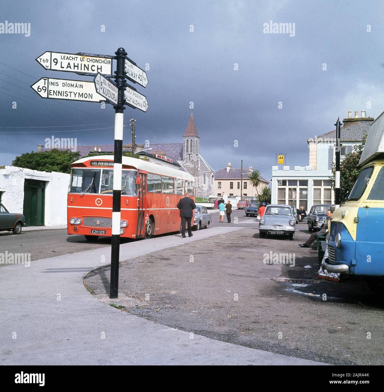 1960s, storico, un autobus pubblico rosso parcheggiata strada presso la città termale di Lisdoonvarna, County Clare, Irlanda, mostrando in lontananza la guglia della chiesa gotica vittoriana romana cattolica revival. Famosa in quest'epoca per il suo festival musicale, è ora un evento di "adattamento musicale", uno dei più grandi d'Europa, che è la principale attrazione turistica per la città. Foto Stock