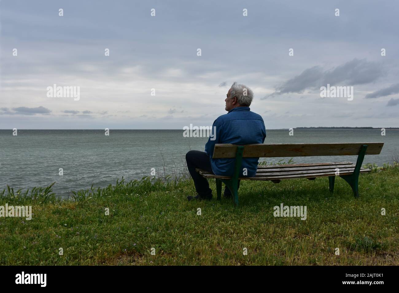 Uomo anziano seduto su una vecchia panca di legno sopra il vedere in un giorno nuvoloso, contemplando il nostalgico con vista sul mare Foto Stock