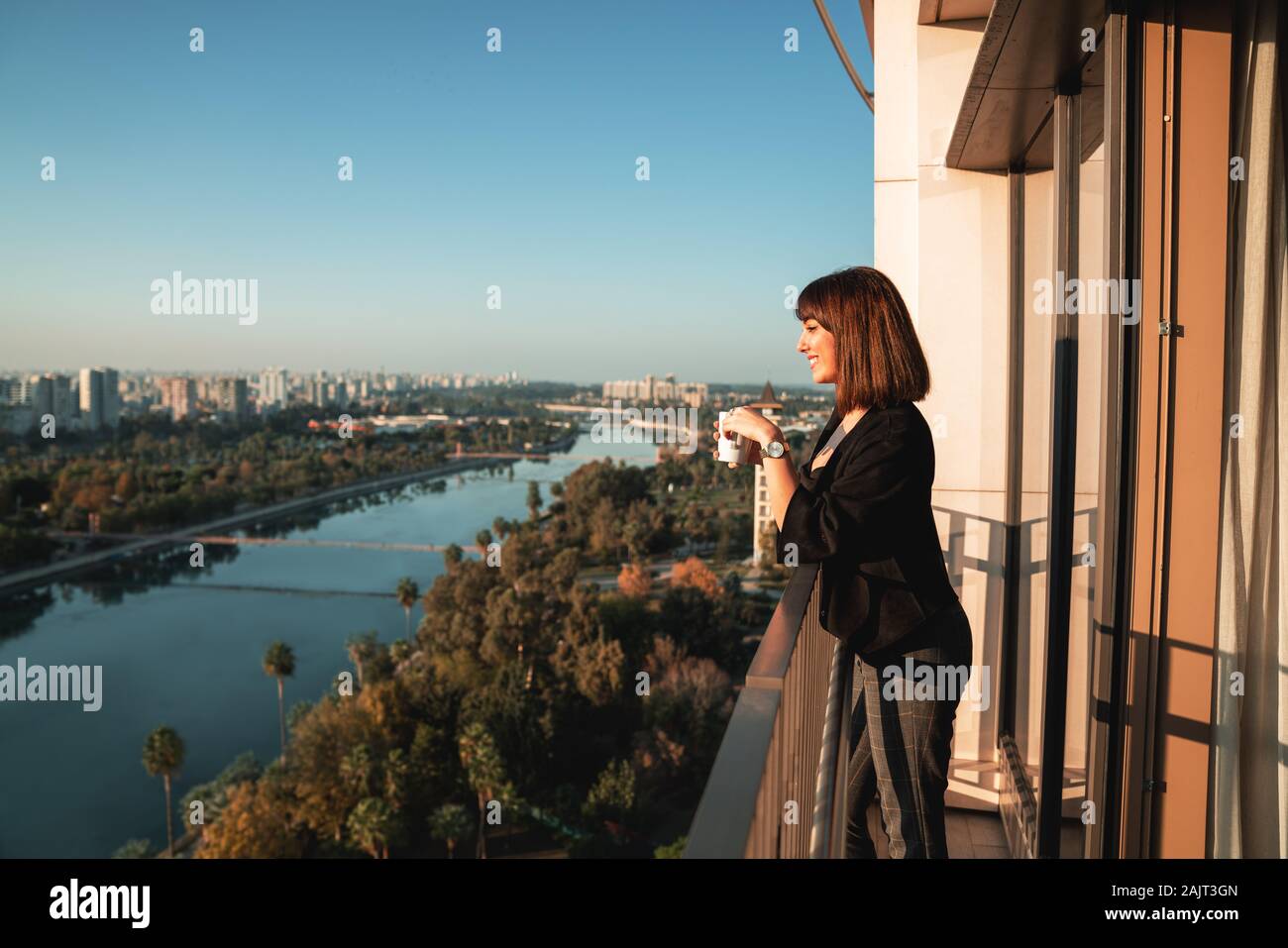 Giovane donna sorridente e bere qualche bevanda e guardando fuori dal balcone. Foto Stock