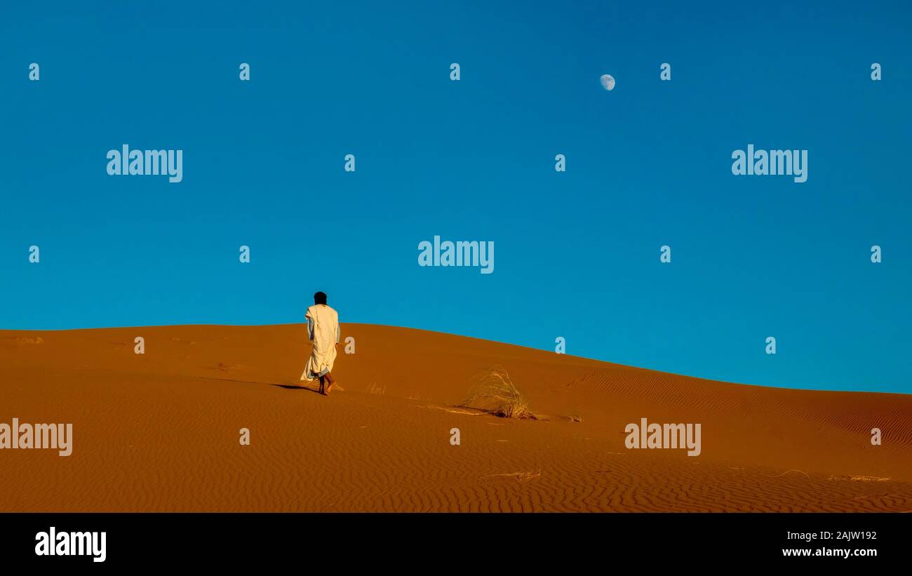 Un marocchino uomo che indossa a lunghezza piena tradizionale abbigliamento nomade, passeggiate a piedi nudi in salita verso il crinale di un rosso dune di sabbia nel deserto del Sahara. Foto Stock