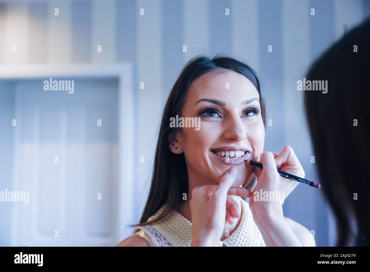 Sorridente giovane ragazza dai capelli neri che guarda in su durante la sessione del trucco da un artista professionale del trucco che disegna una linea di correzione sul suo labbro inferiore Foto Stock