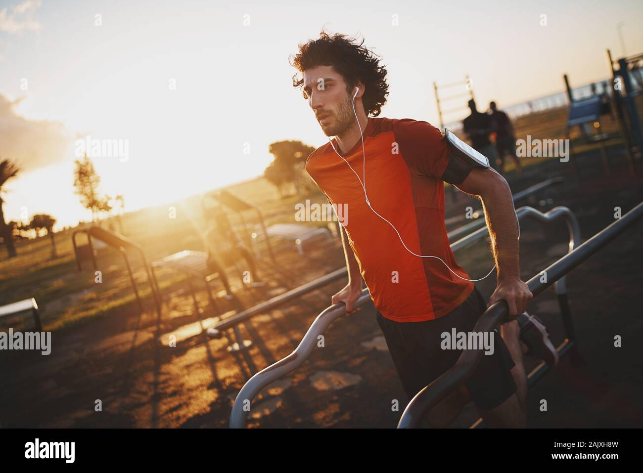Montare il giovane uomo che ascolta musica in cuffia facendo allenamento sul bar nel parco sulla giornata di sole Foto Stock