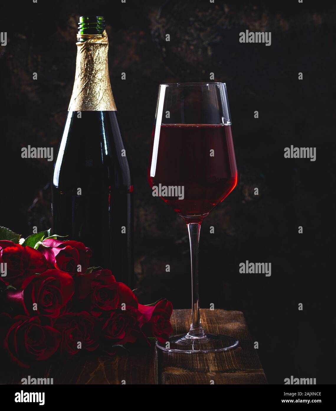 Impostazione scuro di un calice di vino e un mazzo di rose rosse con bottiglia in background su un tavolo di legno Foto Stock