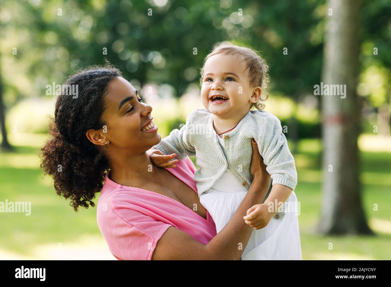 Giovane madre tenendo la sua felice figlia oh mani nel parco. Poco sorridente ragazza che guarda lontano mentre madre portando la sua. Foto Stock