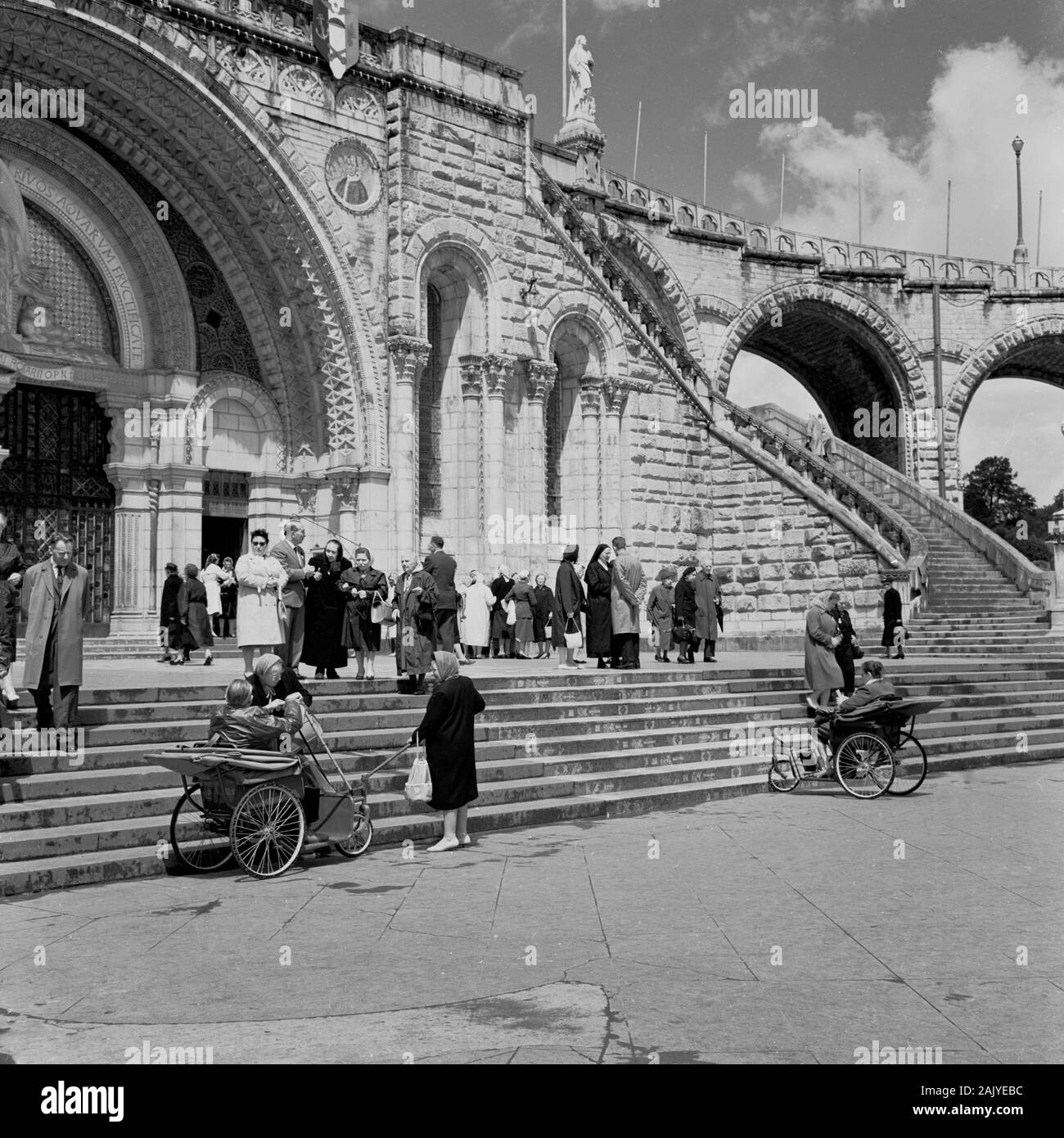 1950s, i visitatori all'ingresso della Basilica del Rosario, Lourdes, Francia, una chiesa cattolica presso il "Santuario della Madonna di Lourdes" o il dominio, un'area di terra che circonda il santuario cattolico (Grotta). Luogo di pellegrinaggio per molti della fede cristiana, specialmente per quelli malati e infermi, come si può vedere nella foto da parte di persone nelle carrozze invalide dell'epoca. Foto Stock