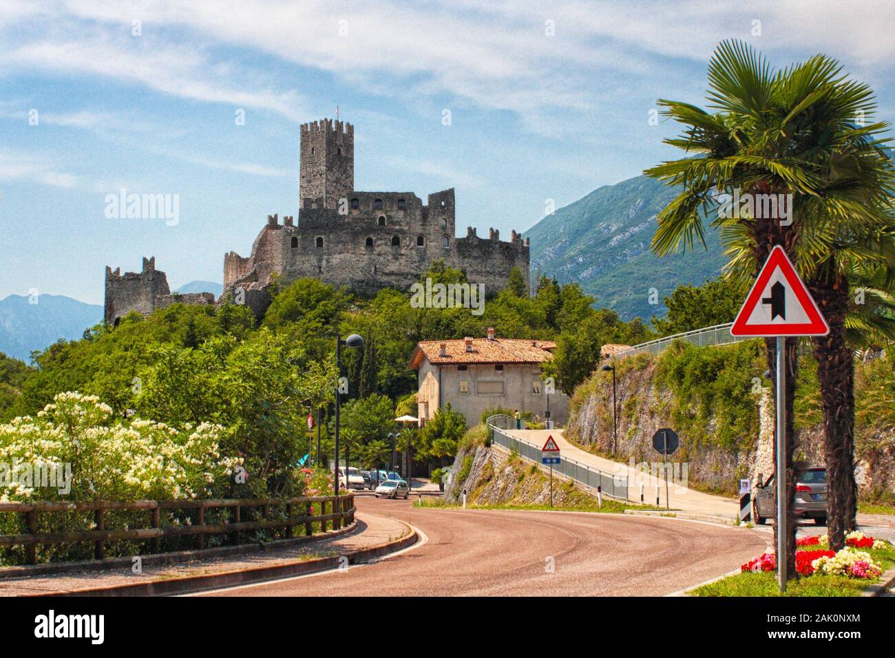 Antico castello con torre in collina. Strada, palme e cespugli in primo piano, Alpi montagne sullo sfondo, Drena, Italia Foto Stock