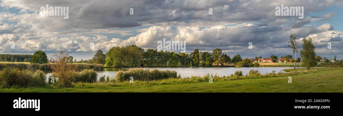 pittoresco paesaggio di campagna con stagno, prato e alberi, in background villaggio e belle nuvole bianche Foto Stock