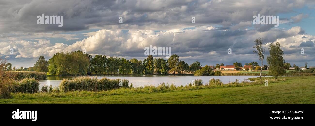 pittoresco paesaggio di campagna con stagno, prato e alberi, in background villaggio e belle nuvole bianche Foto Stock