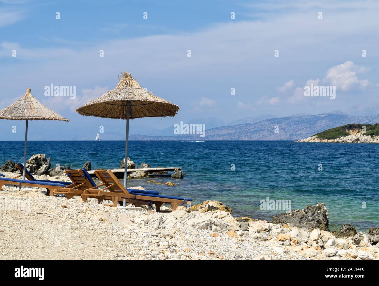 Spiaggia rocciosa sulla riva del Mar Ionio con lettini da mare e ombrelloni di paglia. Il blu e il mare tourquse in background, nessuno. Soleggiata giornata estiva. Ksamil, Al Foto Stock