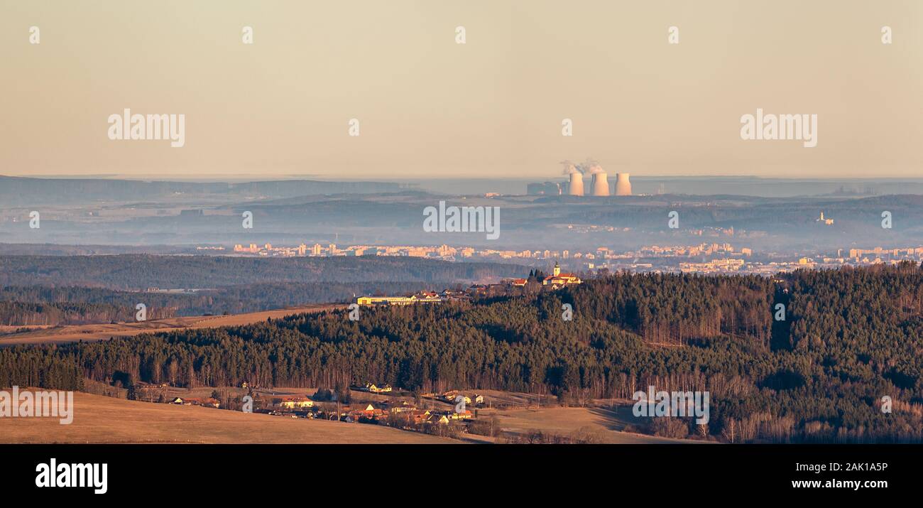 Paesaggio - foresta e collina con la chiesa in primo piano, la città sullo sfondo e le centrali nucleari camini (Temelin, Cechia) all'orizzonte Foto Stock