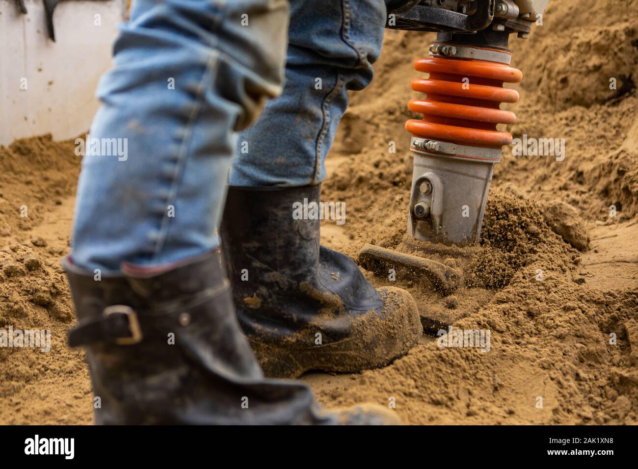 Una chiusura e messa a fuoco selettiva shot sulle gambe di un uomo con una pesante compattatore di massa su un cantiere dopo la posa di fognature, con spazio di copia Foto Stock