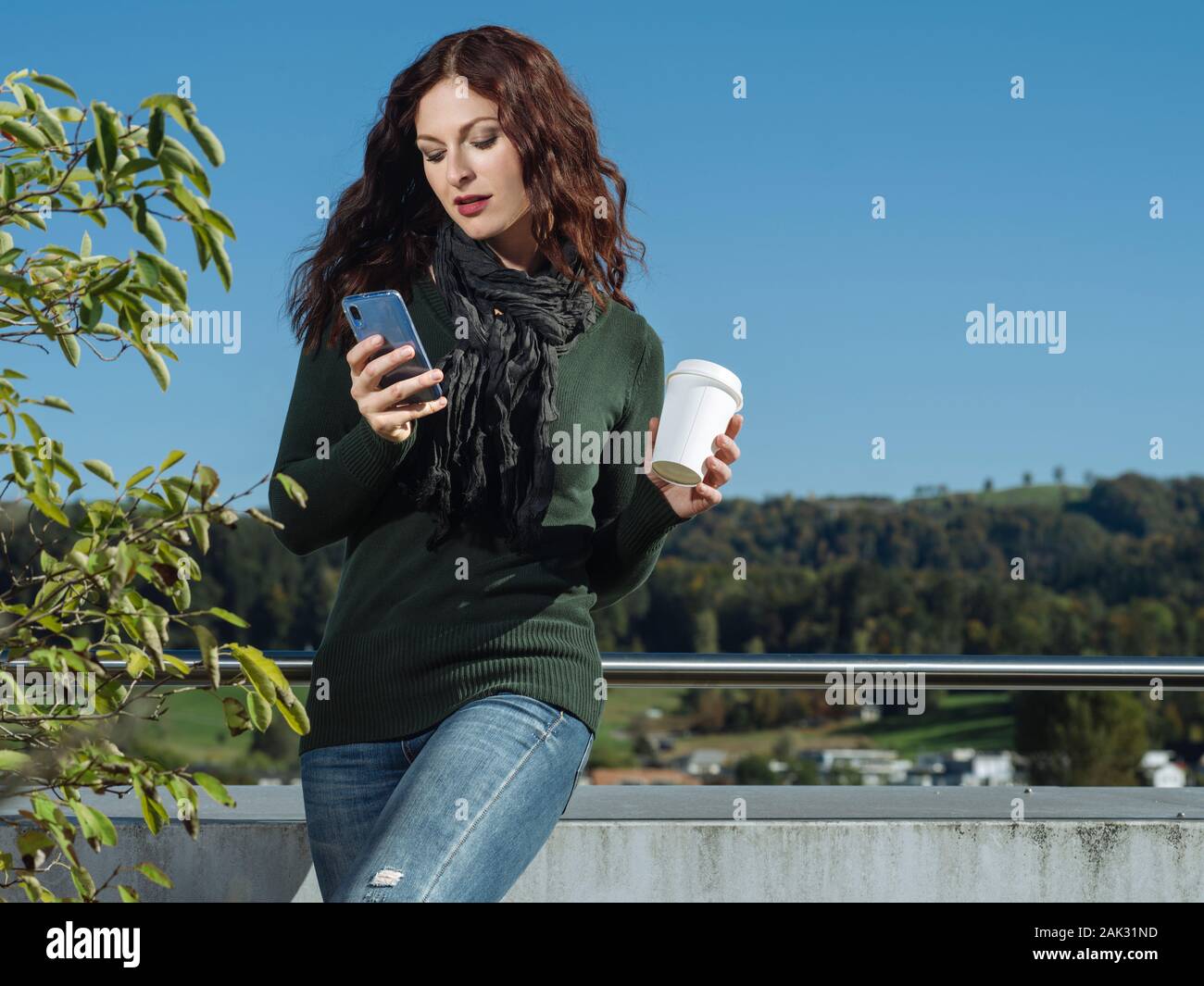 Giovane donna con i capelli rossi utilizzando il suo cellulare e di bere il caffè da una tazza di asporto. Foto Stock