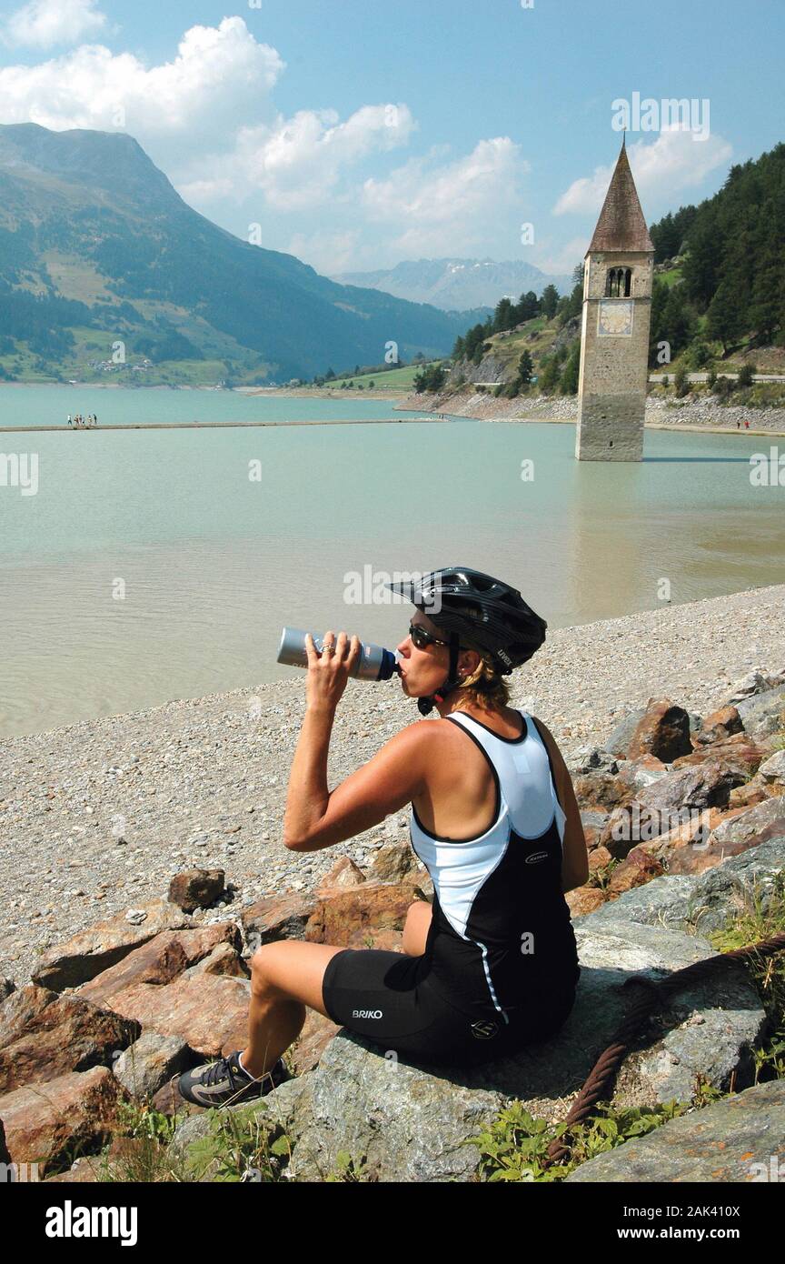 Biken auf der "Via Claudia Augusta" mit einer Trinkpause am Reschensee, Rechts im Bild der Kirchturm von Graun, Südtirol, Italien | Utilizzo di tutto il mondo Foto Stock