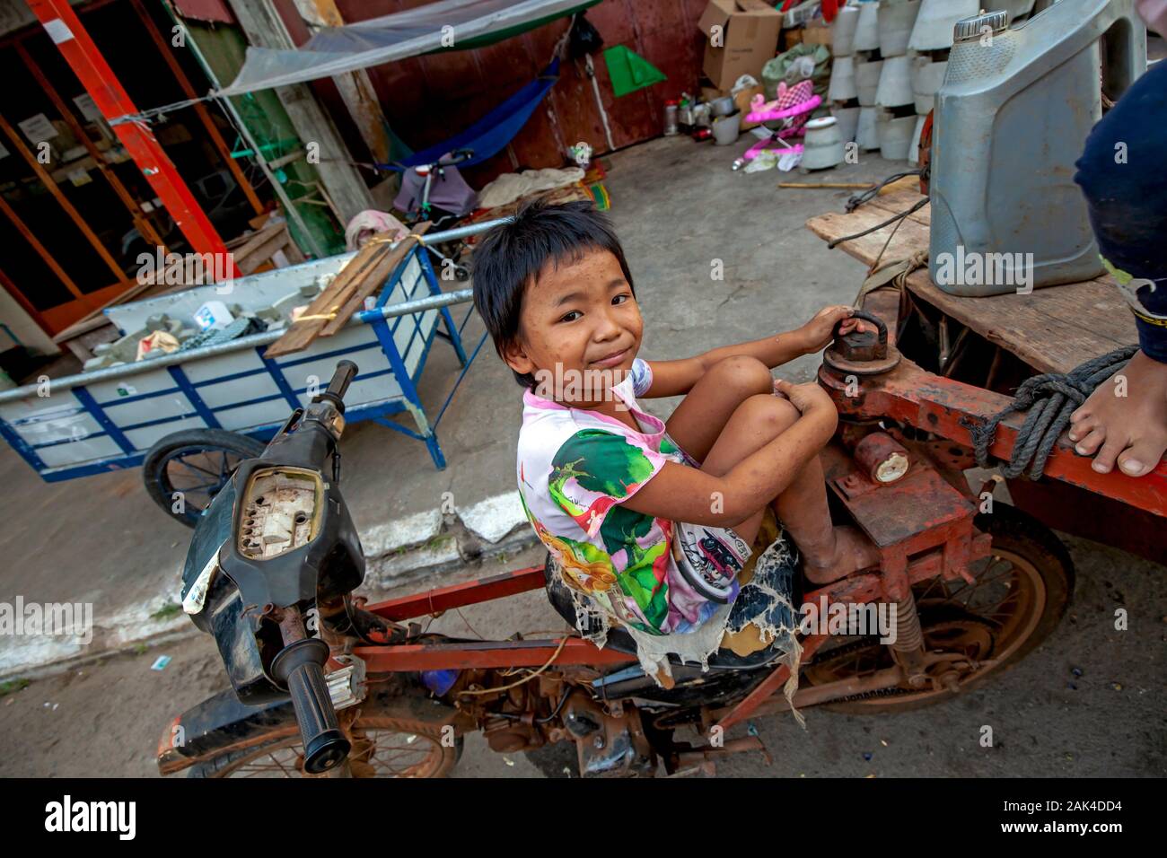 Una povera ragazza la cui famiglia senzatetto purifica per materiale riciclabile siede su un motociclo sedile di fronte al suo rifugio in Kampong Cham, Cambogia. Foto Stock