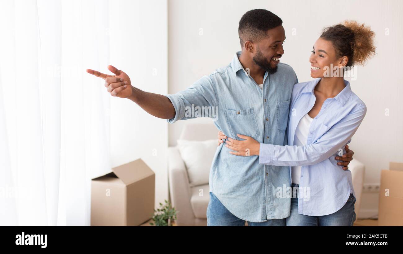 La delocalizzazione. Amare Afro uomo mostra ragazza la vista dalla loro nuova casa abbracciando in piedi in casa propria. Panorama Foto Stock