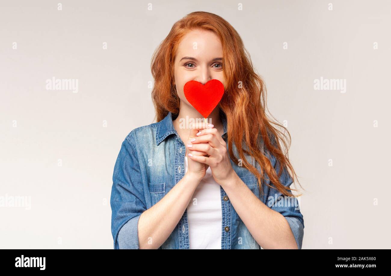 Sono caduto nell'amore. Emozionato redhead teen ragazza che copre la bocca con cuore di carta cercando di non mostrare la sua romantica sentimenti, spazio di copia Foto Stock