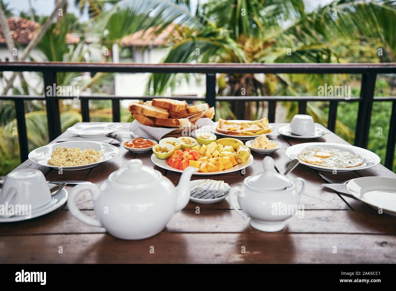 La colazione pronta da mangiare. Uova, frutta tropicale, toasts e tè pentola sul tavolo di legno contro il palm tree. Foto Stock