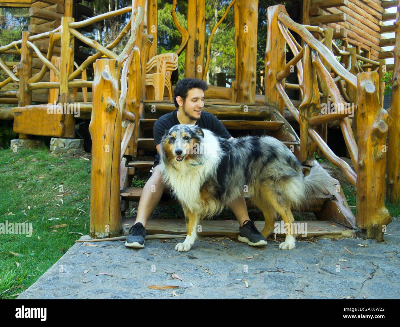 Giovane uomo caucasico petting a tre gambe ruvida Collie cane. L'uomo è seduto su una scala di legno. Amore Animali concetto. Foto Stock