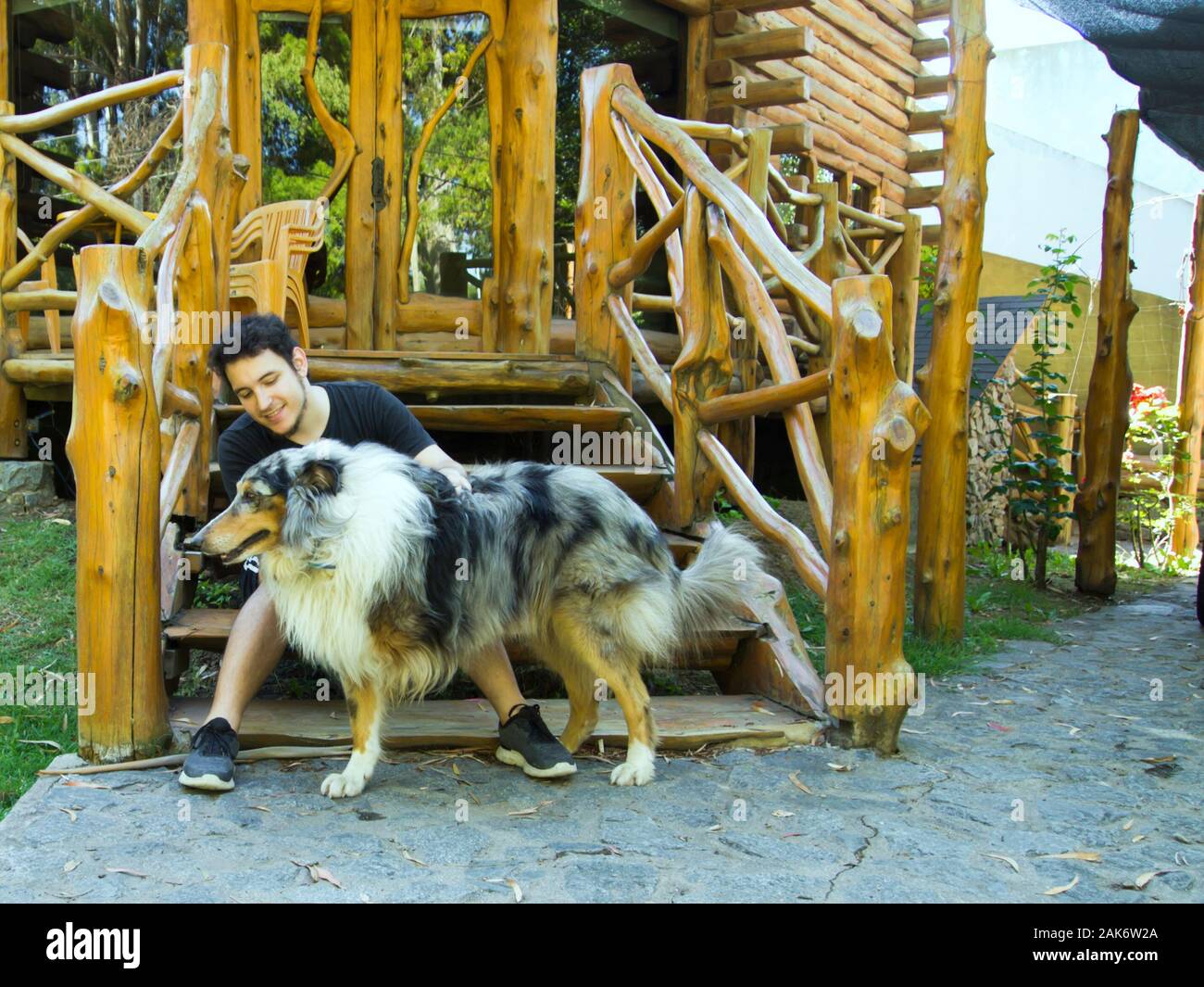 Giovane uomo caucasico petting a tre gambe ruvida Collie cane. L'uomo è seduto su una scala di legno. Amore Animali concetto. Foto Stock
