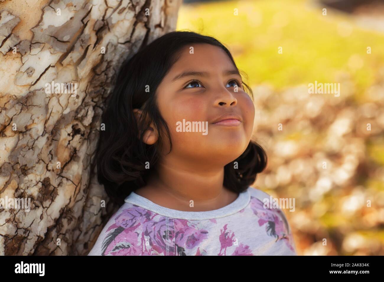 Un latino ragazza con un dolce sorriso, appoggiato su di un albero e guardando il cielo con un senso di speranza. Foto Stock