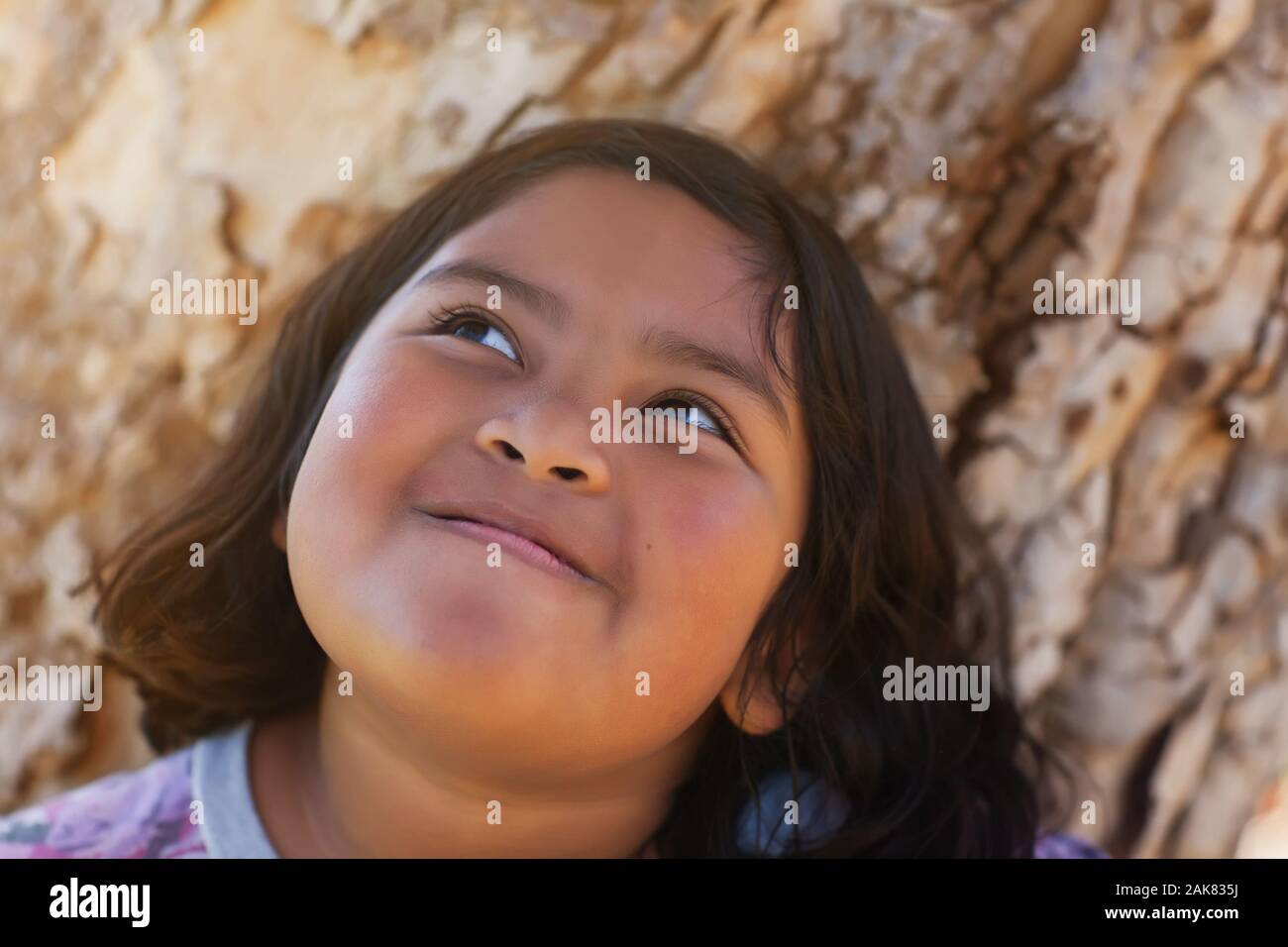 Una giovane ragazza con chubby guance che è piena di gioia e di sorridere mentre in piedi accanto ad un albero e godersi il sole. Foto Stock