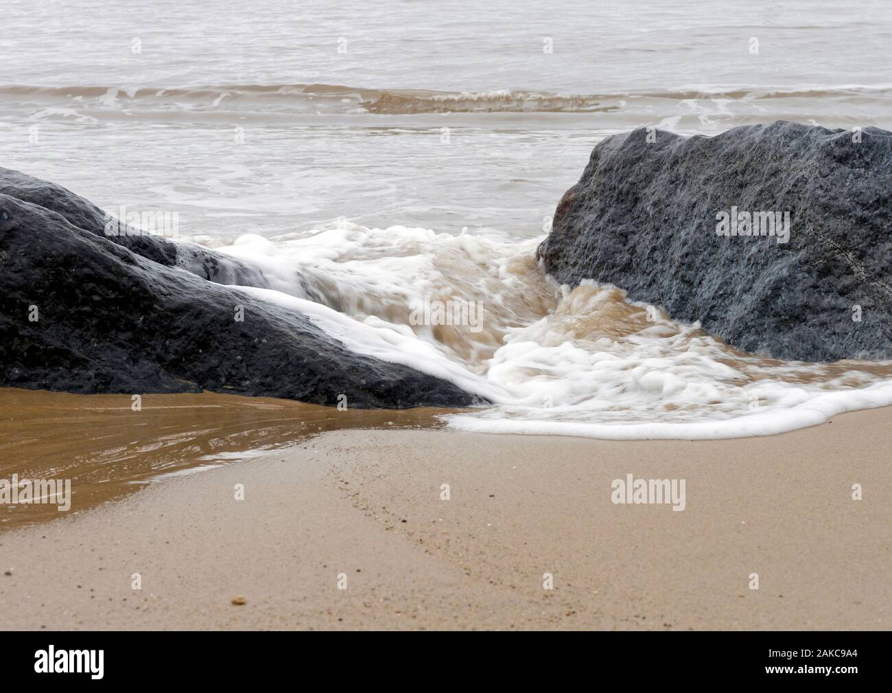 Sabbia, mare e roccia - Una delicata spruzzi d'onda intorno a grandi rocce collocato su una spiaggia di Norfolk come parte delle difese del mare. Foto Stock