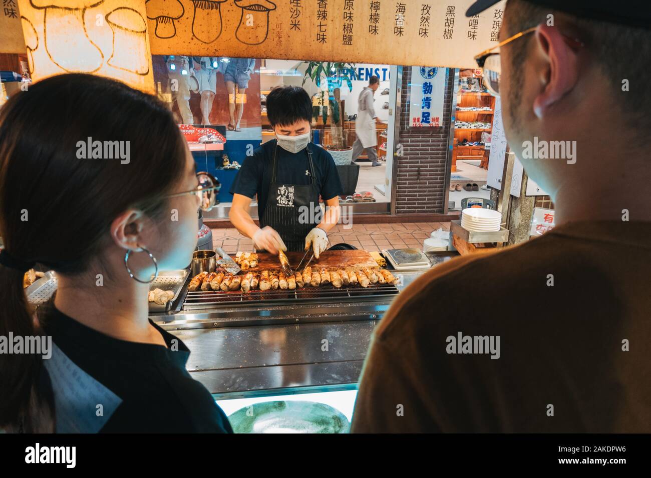 Due clienti guardano come un venditore prepara i funghi di ostriche alla griglia al mercato notturno di Shi Lin, Taipei, Taiwan Foto Stock