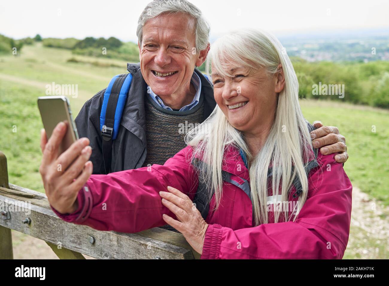Sorridente coppia Senior escursionismo in campagna in piedi da Gate e tenendo Selfie sul telefono cellulare Foto Stock