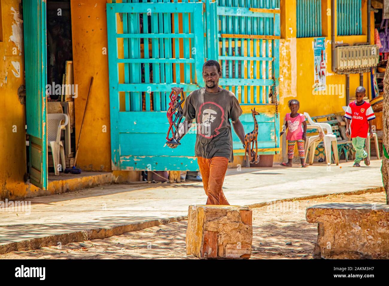 Isola di Goree, Senegal- 22 aprile 2019: l'uomo non identificato a piedi lungo una strada di sabbia in una piccola città e trasportare un legno scolpito figurine. Foto Stock