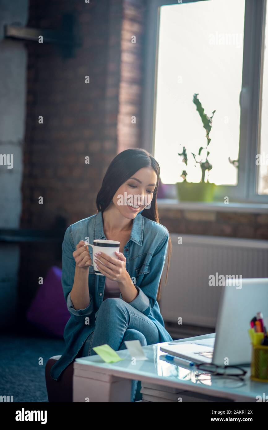 Bella donna positiva di bere il caffè sul posto di lavoro Foto Stock