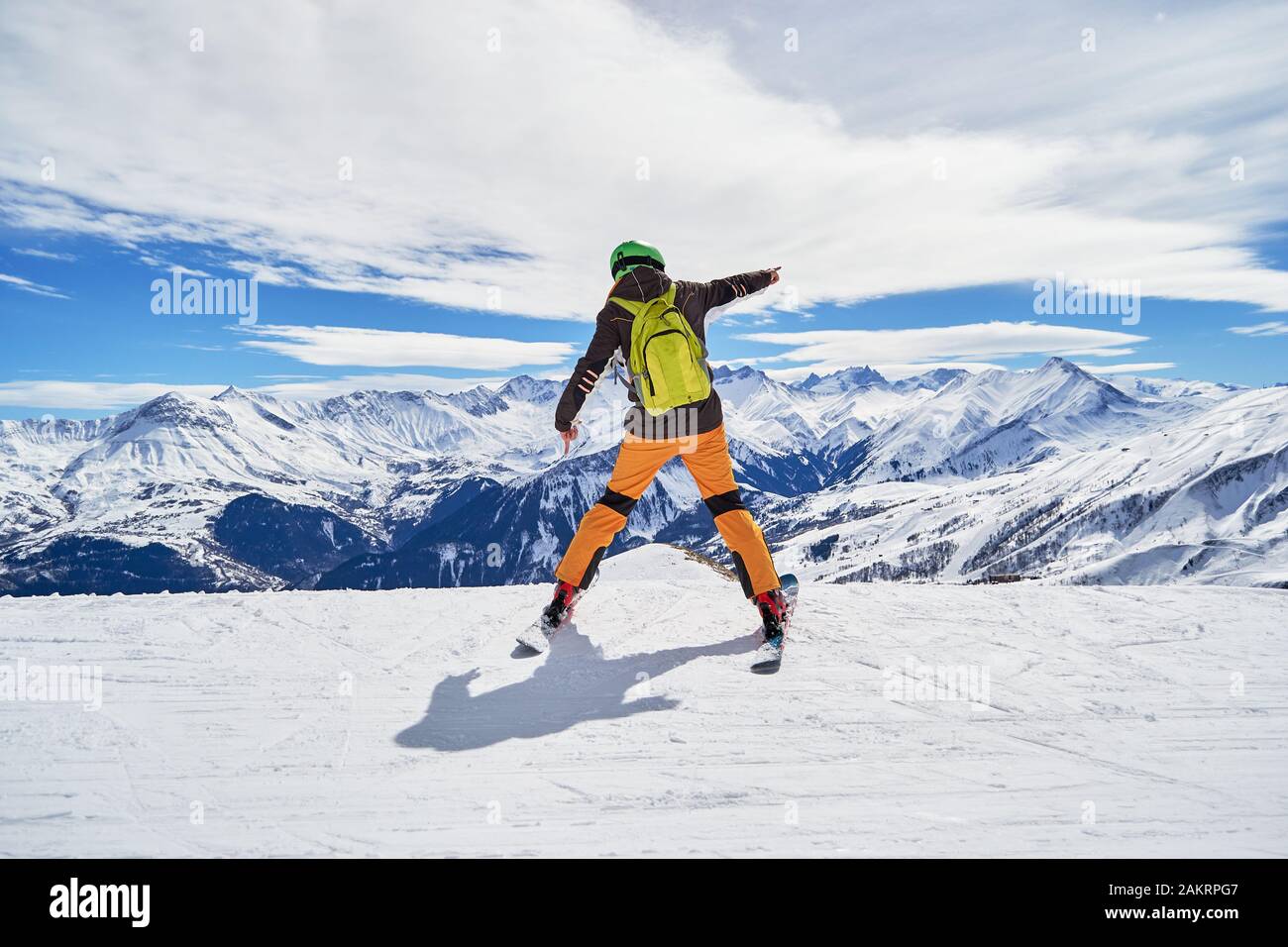 Lo sciatore entusiasta indossa abiti colorati e uno zaino verde, che si posa su una pista da sci nella località sciistica di Les Sybelles, con le vette delle Alpi francesi sul retro Foto Stock
