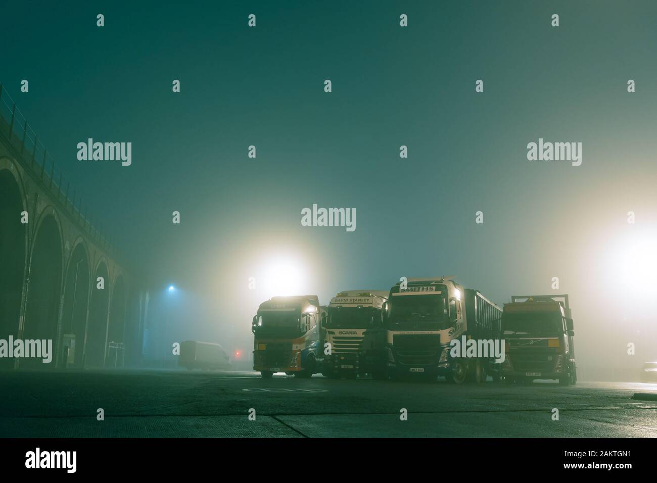 Quattro camion parcheggiato in un parcheggio, con le luci di strada dietro su un moody, atmosferici foggy inverni di notte. Foto Stock