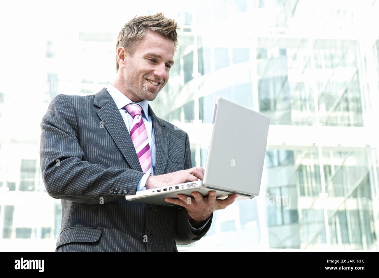 Uomo d'affari sorridente in piedi all'aperto digitando sul suo computer portatile di fronte allo sfondo di moderne torri di uffici in vetro Foto Stock